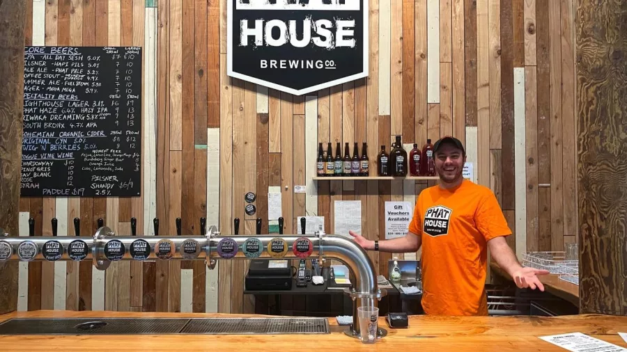 Bartender behind the tap line at Phat House Brewing bar with a wall of craft beer taps and bottles