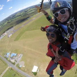 Tandem skydivers descending toward the Bay of Islands landing zone flashing peace signs and smiling