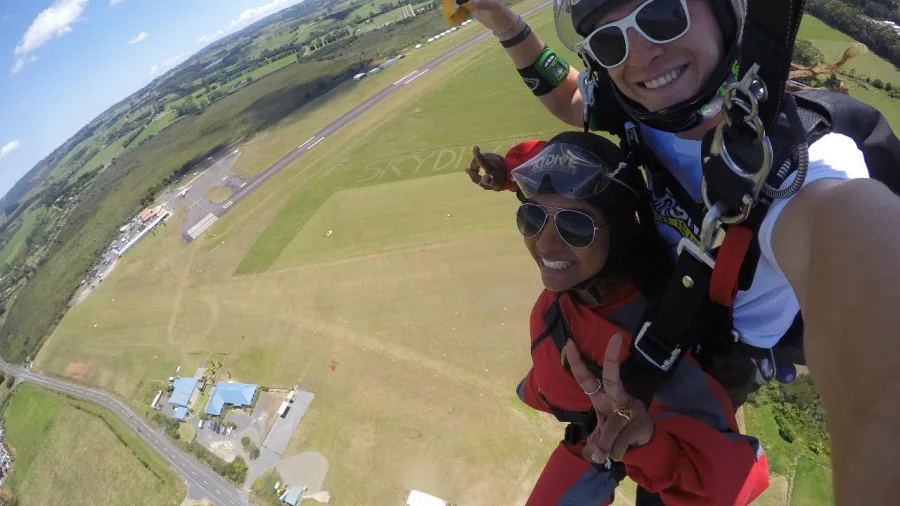 Tandem skydivers descending toward the Bay of Islands landing zone flashing peace signs and smiling