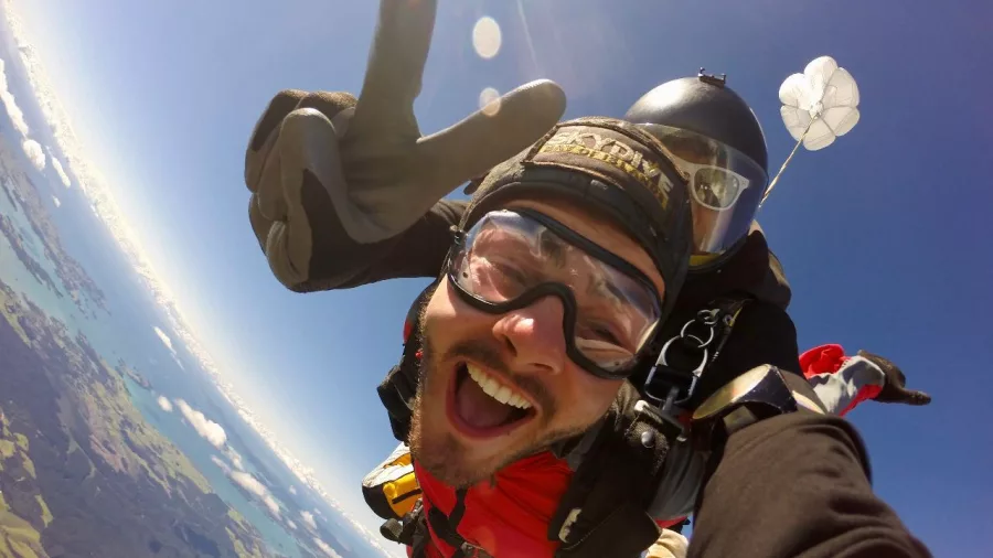 Tandem skydivers smiling with a peace sign mid-freefall over Northland with clear skies and landscape below