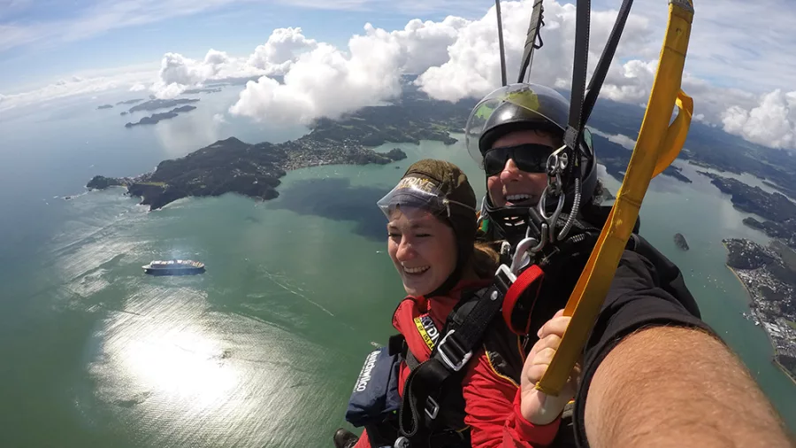 Tandem skydivers descending under parachute with coastal views of the Bay of Islands and surrounding waters below.