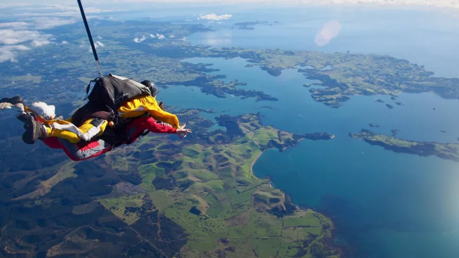 Tandem skydivers freefalling above the Bay of Islands, with clear views of the coastline, ocean, and islands below.