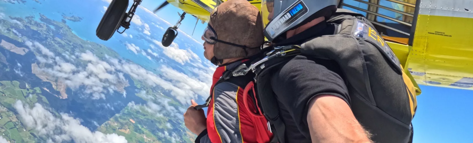 Tandem skydivers mid-air just after exiting a yellow aircraft, high above the North Island with cloud patterns below