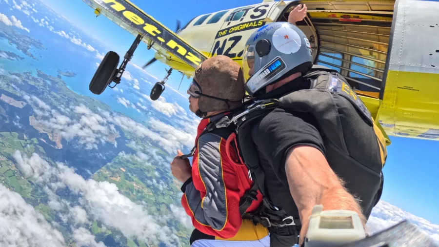 Tandem skydivers mid-air just after exiting a yellow aircraft, high above the North Island with cloud patterns below