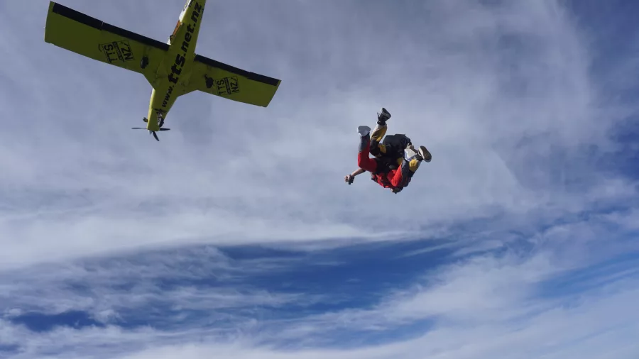 Tandem skydivers tumbling in freefall moments after jumping from a yellow aircraft over New Zealand