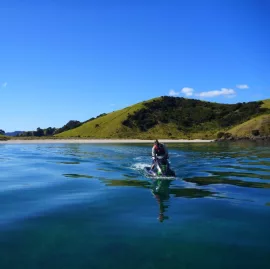 A person riding a jet ski toward a quiet beach surrounded by green hills in the Bay of Islands