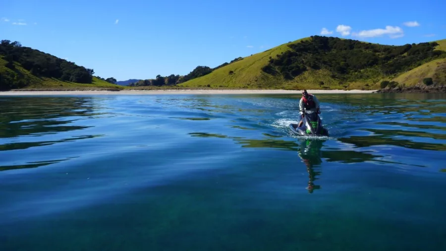 A person riding a jet ski toward a quiet beach surrounded by green hills in the Bay of Islands