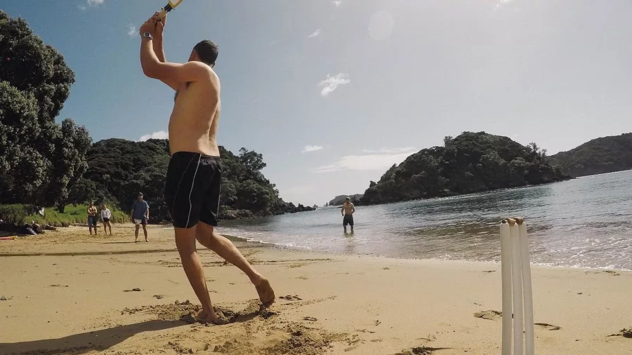 Man playing beach cricket with a group on the shore during The Rock Adventure Cruise in the Bay of Islands