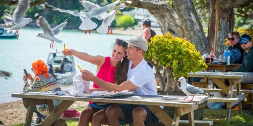 Couple enjoying fish and chips by the waterfront at Kororāreka Bay in Russell, Northland, with seagulls flying overhead