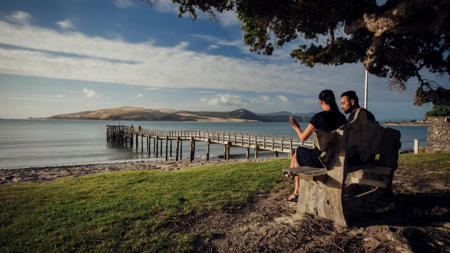 Couple relaxing on a wooden bench with views of Ōmāpere Wharf and Hokianga Harbour