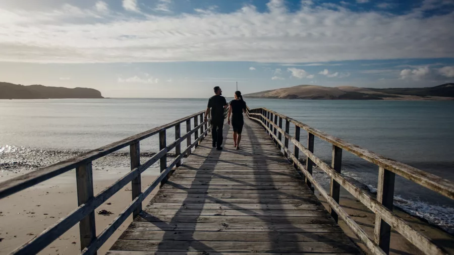 Couple walking hand-in-hand along Ōmāpere Wharf with the sand dunes in the distance
