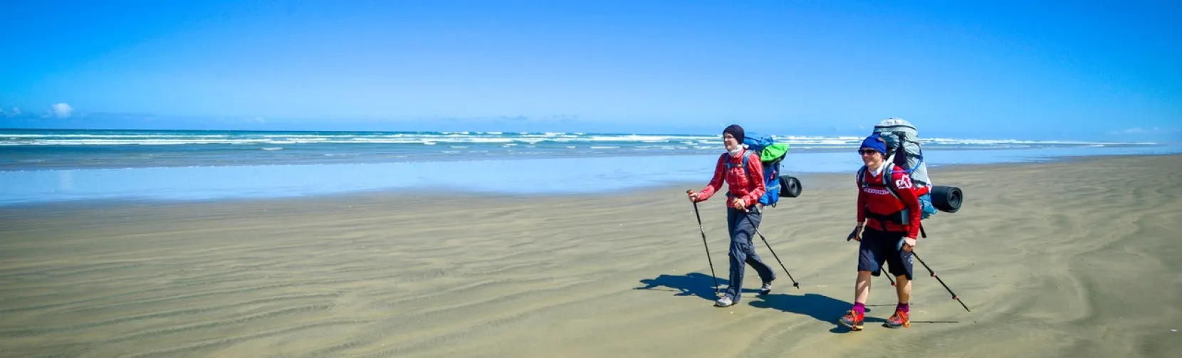 Two hikers with backpacks walking along the wide sands of Ninety Mile Beach in Northland, New Zealand
