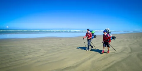 Two hikers with backpacks walking along the wide sands of Ninety Mile Beach in Northland, New Zealand