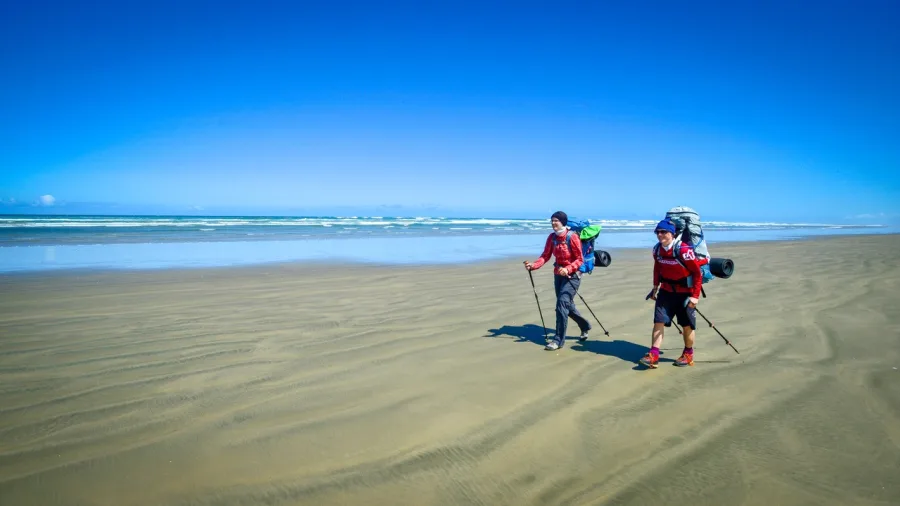 Two hikers with backpacks walking along the wide sands of Ninety Mile Beach in Northland, New Zealand