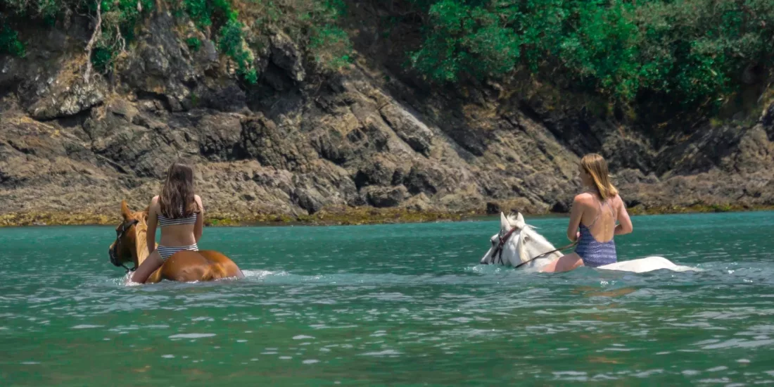 Two riders bareback on horses swimming in clear blue waters near rocky cliffs
