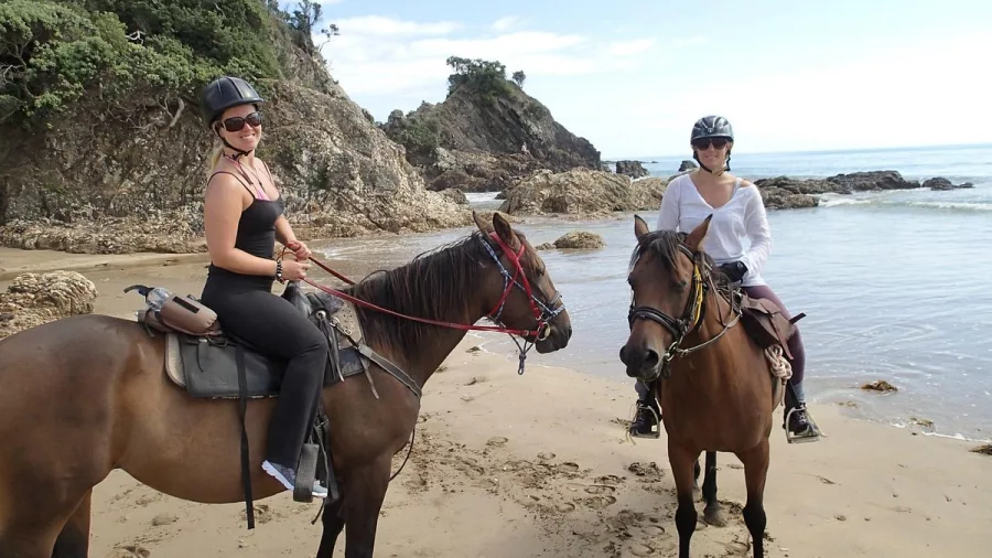Two women on horseback smiling during a coastal trek in Matapōuri, New Zealand