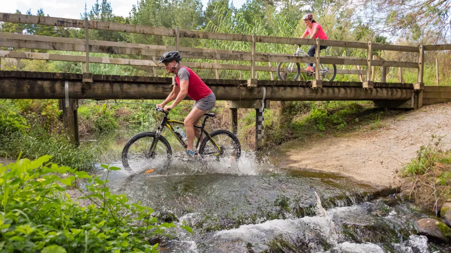 Cyclist splashing through a shallow stream crossing with a wooden bridge in the background