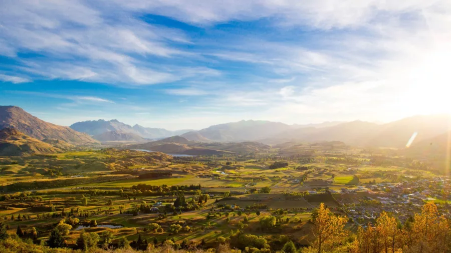Panoramic view from Tobins Track in Arrowtown during autumn, with sunlit mountains, rolling hills, and vibrant foliage.