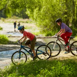 Two children cycling near the Arrow River in Arrowtown, New Zealand, with families relaxing by the riverside in the background.