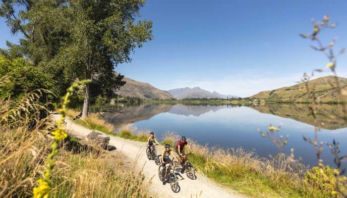 Cyclists riding along the lakeside trail at Lake Hayes near Arrowtown, New Zealand, with mountain reflections on a clear day.