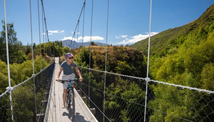 Cyclist crossing the Discovery Bridge on the Arrow River Bridges Trail in Arrowtown, New Zealand, with mountain and forest views.