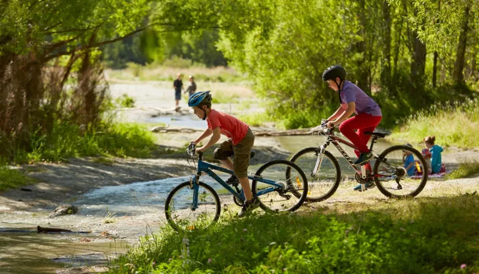 Two children cycling near the Arrow River in Arrowtown, New Zealand, with families relaxing by the riverside in the background.