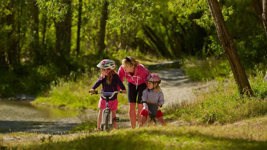 Family cycling together along Arrowtown River Trail in New Zealand on a sunny day.