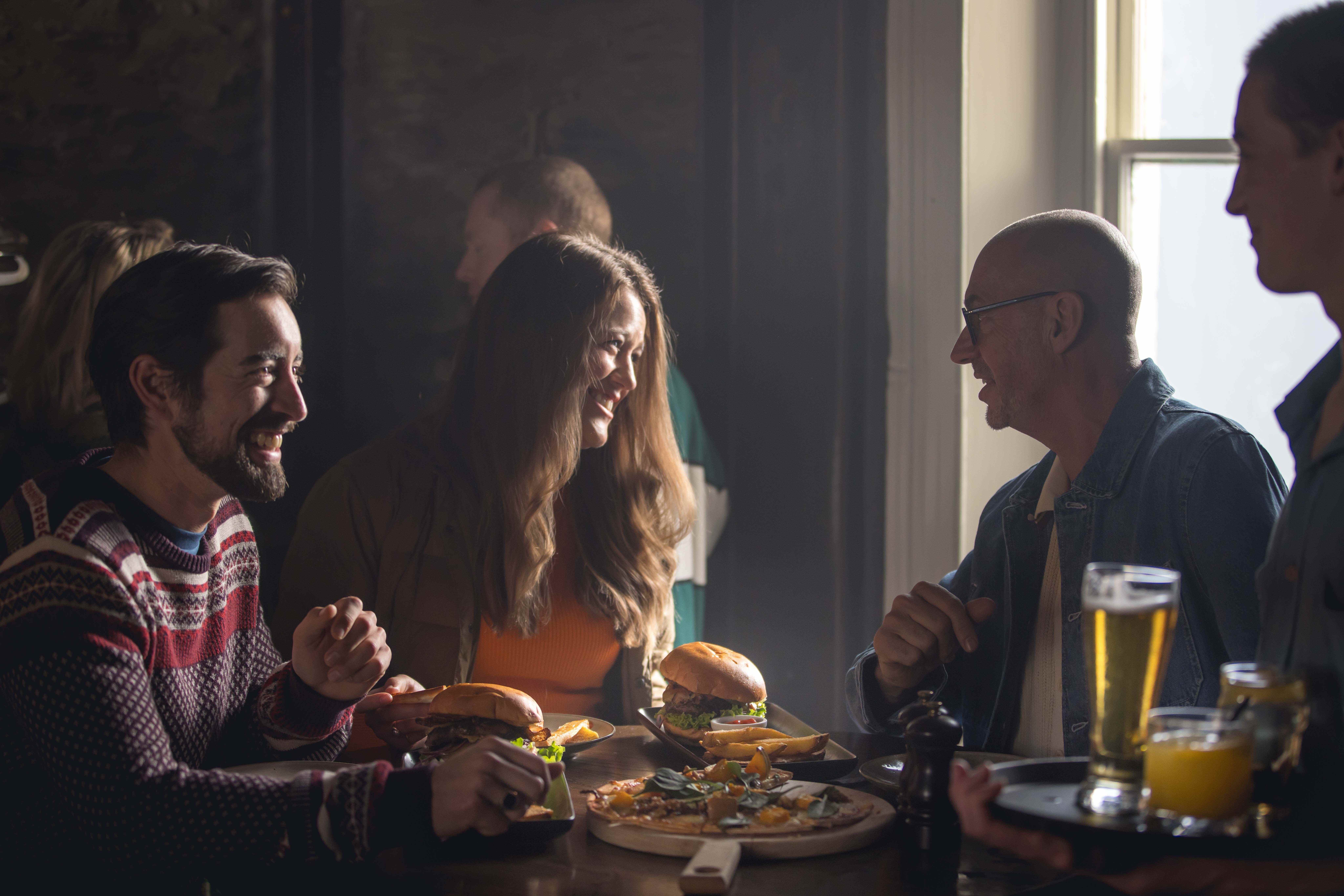A group of people dining indoors in Arrowtown near Queenstown, New Zealand, with burgers, pizza, drinks, and warm window light in a relaxed restaurant setting. Photo by Miles Holden.