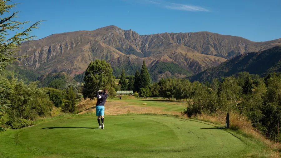 Golfer teeing off at Arrowtown Golf Club with mountain views in the background, New Zealand.
