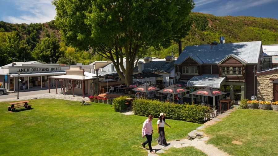 Buckingham Green in Arrowtown, New Zealand, with the historic Bendix Stables built in 1873, outdoor dining areas, and views of surrounding hills.
