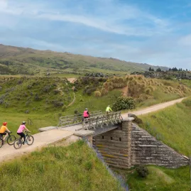 Rural bridge and riders on Otago Central Rail Trail through open farmland
