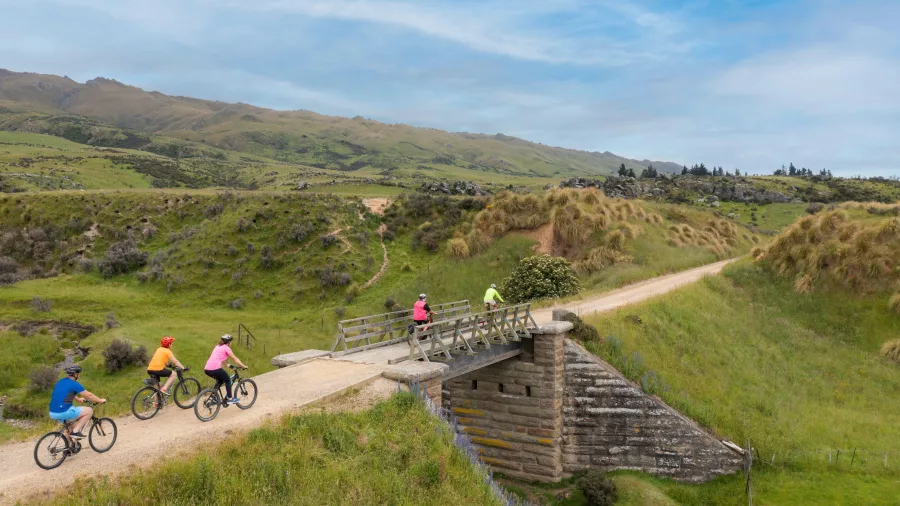 Rural bridge and riders on Otago Central Rail Trail through open farmland