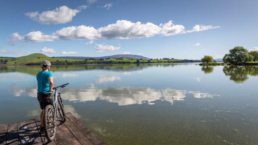 Cyclist standing with bike on a lakeside jetty overlooking the still waters of Lake Waihola on the Clutha Gold Trail near Dunedin, New Zealand