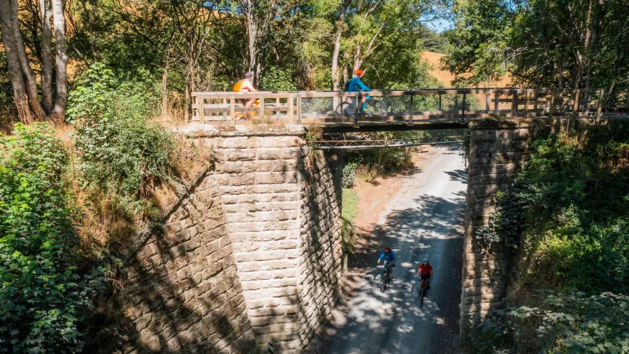 Cyclists crossing and riding beneath a restored heritage bridge on the Clutha Gold Trail between Lawrence and Milton in Central Otago, New Zealand