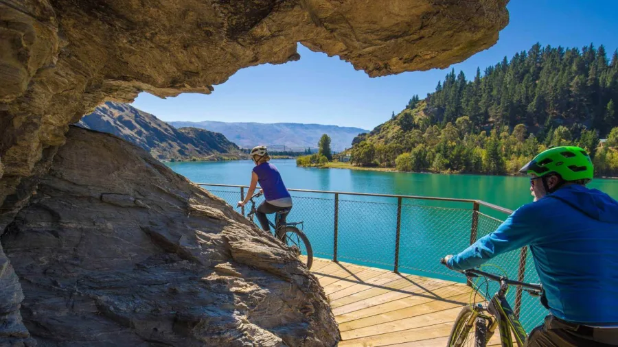 Cyclists riding along a boardwalk built into the cliffs above turquoise waters on the Lake Dunstan Trail.