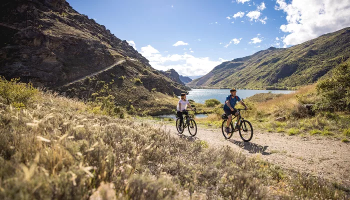 Cyclists riding on a scenic lakeside section of the Lake Dunstan Trail in Central Otago, New Zealand