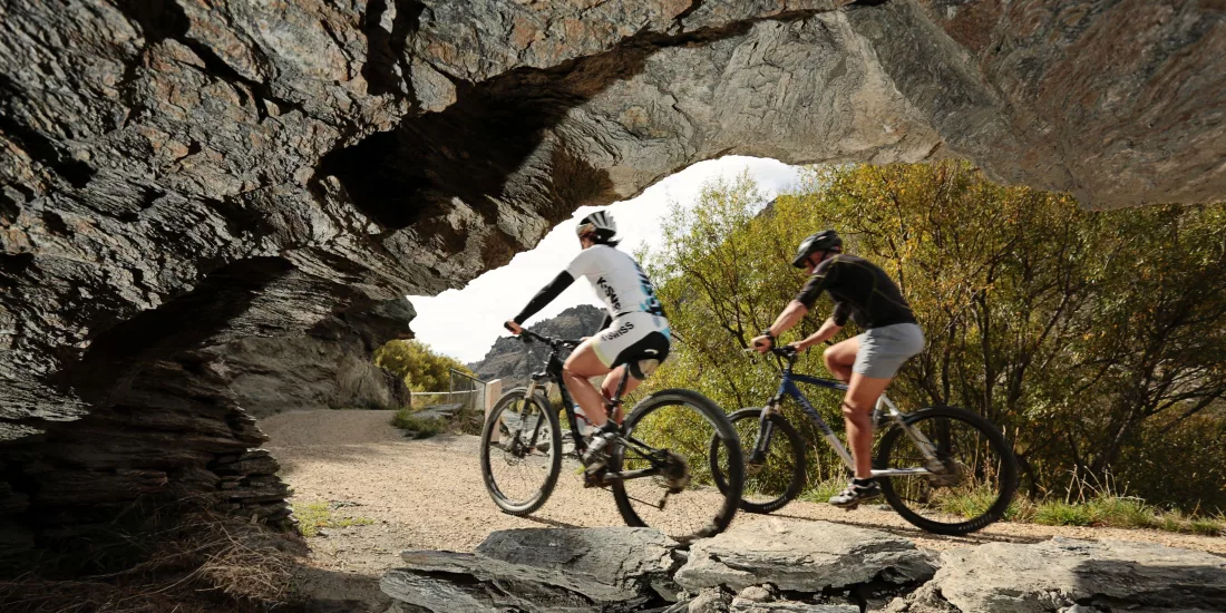Two cyclists ride under a natural rock arch on the Roxburgh Gorge Cycle Trail near Alexandra in Central Otago, New Zealand