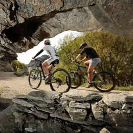 Two cyclists ride under a natural rock arch on the Roxburgh Gorge Cycle Trail near Alexandra in Central Otago, New Zealand