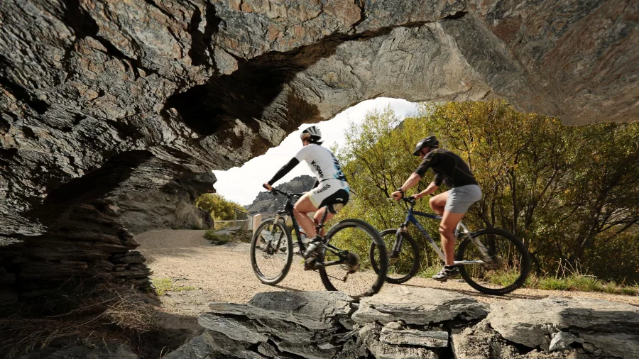 Two cyclists ride under a natural rock arch on the Roxburgh Gorge Cycle Trail near Alexandra in Central Otago, New Zealand