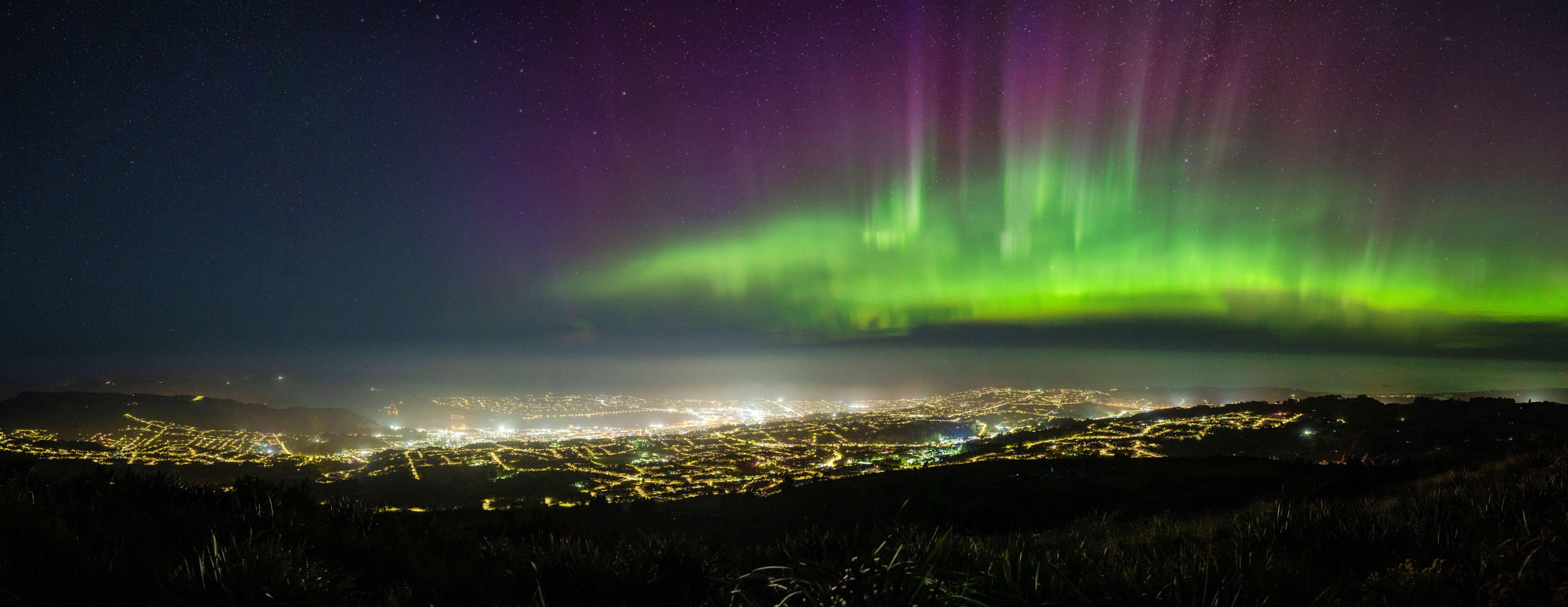 Aurora Australis glowing in green and purple hues above the city lights of Dunedin, South Island New Zealand