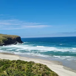 Waves rolling into Smails Beach with rugged coastal cliffs and green hills in Dunedin, New Zealand.