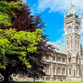 Iconic clocktower building of the University of Otago, surrounded by leafy trees and blue sky.