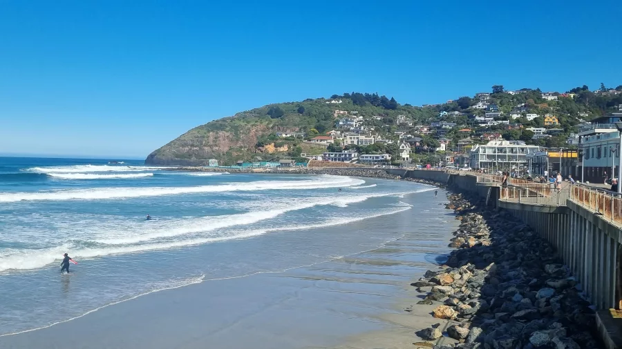 View of the St Clair Esplanade lined with cafés, shops, and surfers catching waves.