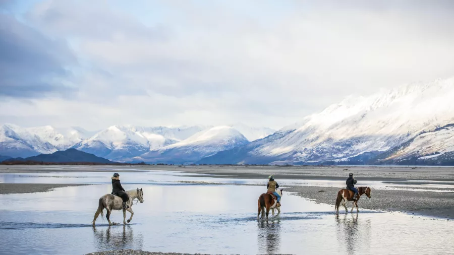 Horse riders crossing shallow water with snowy mountains in the background in Glenorchy.