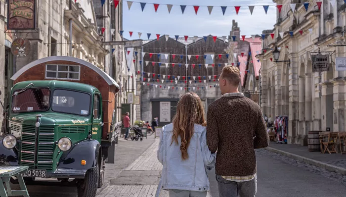Couple walking through Oamaru Victorian Precinct in Waitaki, New Zealand, past heritage buildings and a vintage green truck.