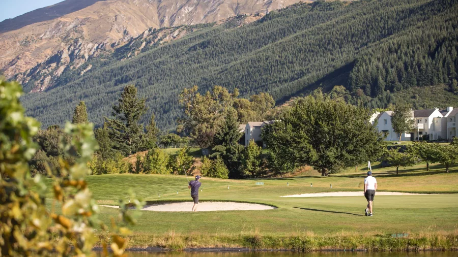Golfers playing near a bunker at Millbrook Resort in Arrowtown with alpine hills and villas in the background