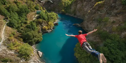 A person in a red shirt mid-air during a bungy jump off the Kawarau Bridge over the turquoise Kawarau River in Queenstown.