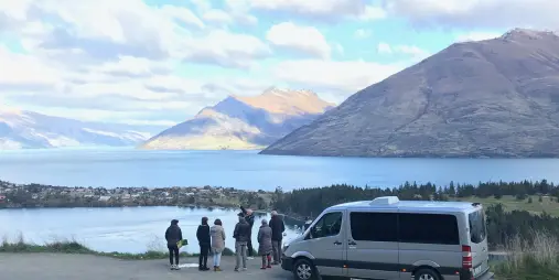 Tour group standing beside a van at Hensman Road Lookout, overlooking Lake Whakatipu and the Remarkables