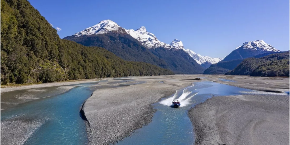 Aerial view of Dart River jet boat travelling through braided river channels surrounded by native forest and mountains.