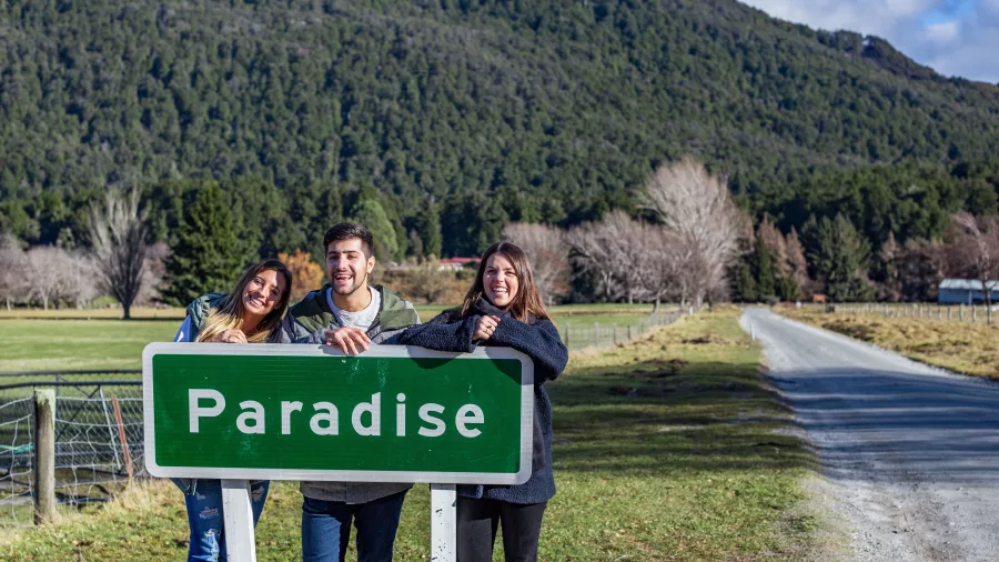 Tour guests posing by the iconic Paradise sign near Glenorchy with forested hills in the background